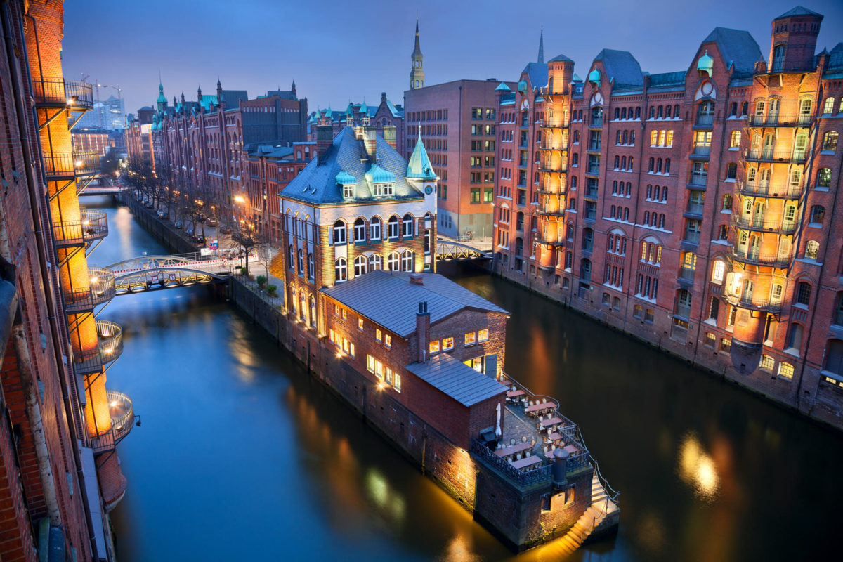 Picture taken from the top, and angled toward the left. The time of dusk can be seen in the sky, lights from the buildings are golden bright. The picture shows a canal at the bottom, it is surrounded by tall buildings of European architecture. The start of the canal's division is visible, it is divided in two parts, the center dividing the two parts expands as there is a building structure that becomes broader. This is in the German city of Hamburg.